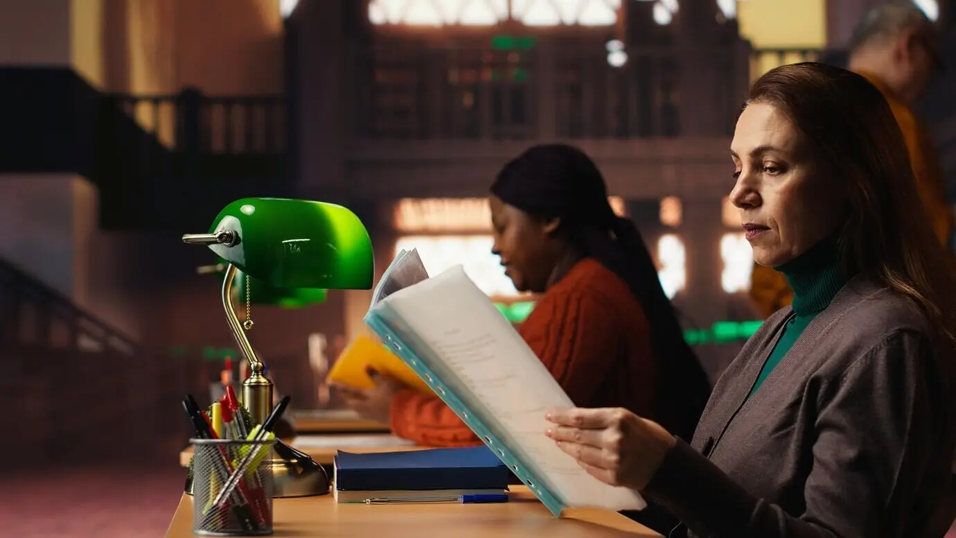 Halk kütüphanesi ortamında, yumuşak aydınlatma altında ders çalışan yaşlı bir kadın.